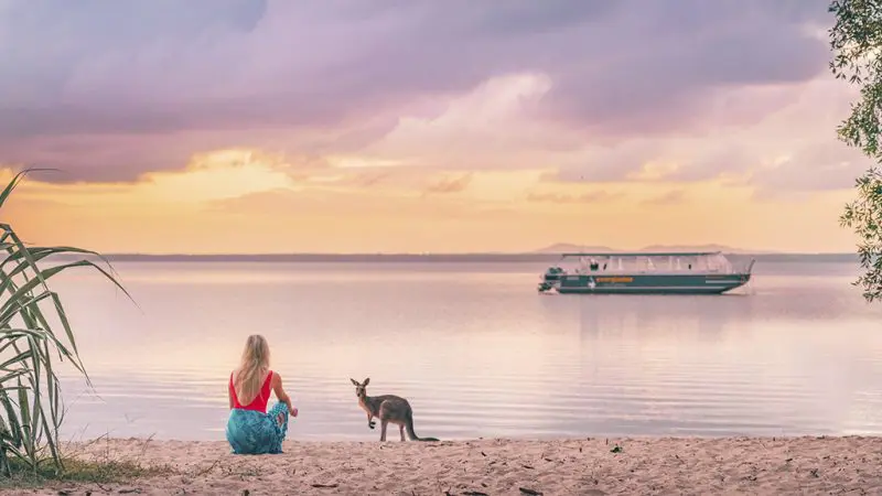 Woman unwinds on sunlit Noosa beach at sunset, kangaroo nearby, highlighting Half Day Noosa Everglades Serenity Cruise experience.