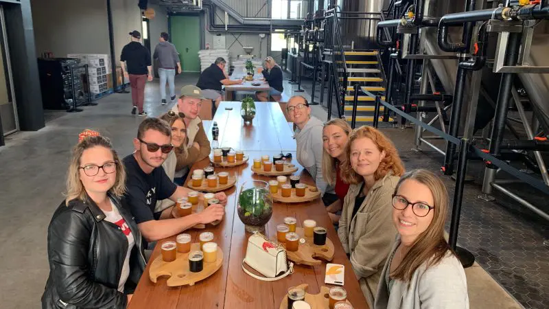 Smiling group savouring assorted beer flights at a long table during a full day brewery and distillery tour inside a vibrant brewery.