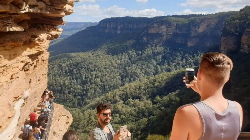 Tourists take photos on a Blue Mountains Day Tour with scenic waterfall walk and group lunch, overlooking lush forested valley views.