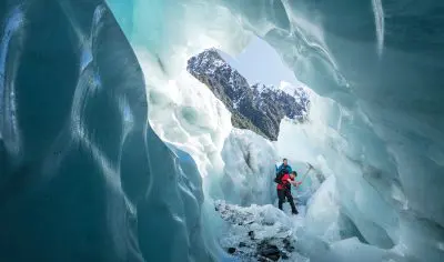 Adventurers in winter gear explore a dazzling blue ice cave on Franz Josef Glacier, snowy mountain peaks visible through the cave entrance.