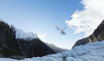 Helicopter glacier tour soaring above snowy Franz Josef Glacier and rugged peaks under a vibrant blue, cloud-streaked sky in New Zealand.