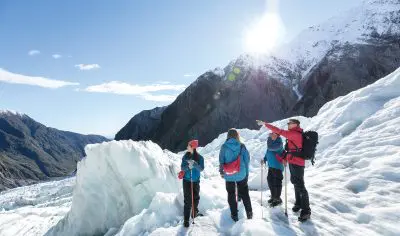 Four hikers in winter clothing explore Franz Josef Glacier under bright sun, with one pointing towards snowy peaks during a scenic heli-hike.