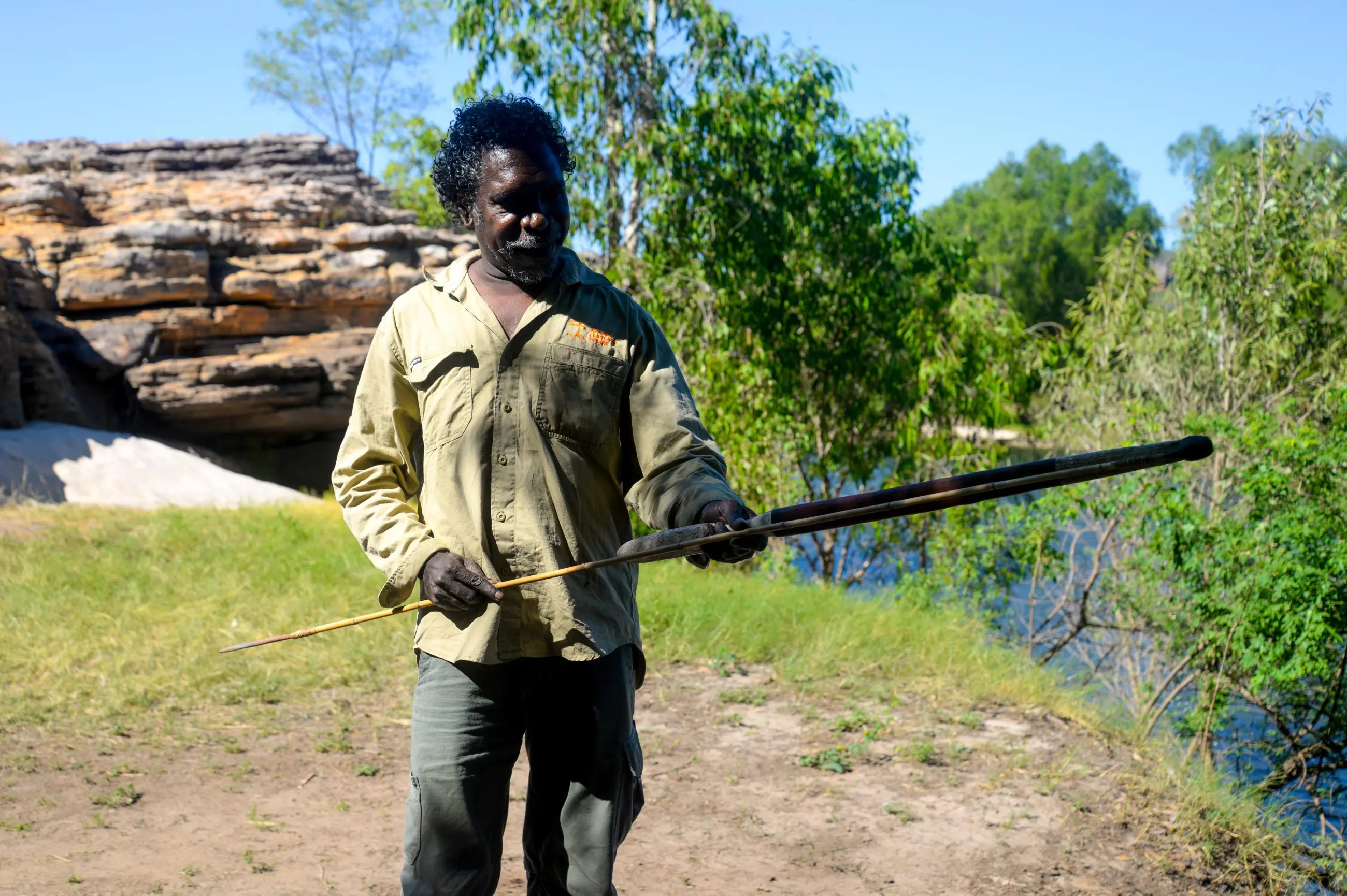 Experience an authentic Kakadu Arnhem Land Tour: a man outdoors demonstrates traditional spear skills amid lush trees and rugged rocks.