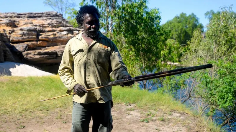 Experience an authentic Kakadu Arnhem Land Tour: a man outdoors demonstrates traditional spear skills amid lush trees and rugged rocks.