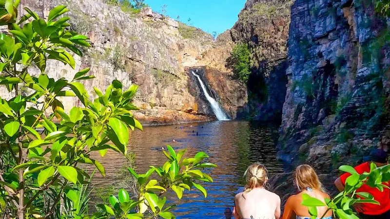 Two women relax beside a rocky natural pool, admiring a stunning waterfall on their Kakadu Arnhem Land Tour under a vibrant blue sky.