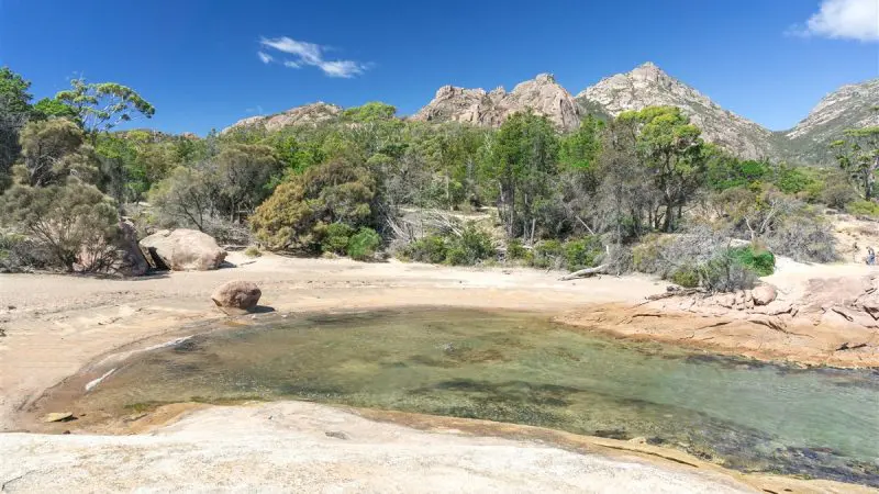 Crystal-clear shallow pool with rocky shoreline, lush trees, and majestic mountains on Wineglass Bay Full Day Tour via Richmond, Tasmania.