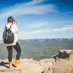 Adventurer photographing breathtaking Blue Mountains scenery from a cliff on an Uncharted Tour, blue sky in the background.