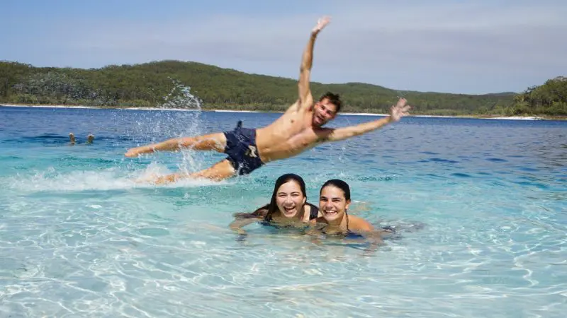 Two women enjoy crystal-clear water while a man leaps in, splashing, on a guided 3 Day K'gari Adventure tour from Rainbow Beach.