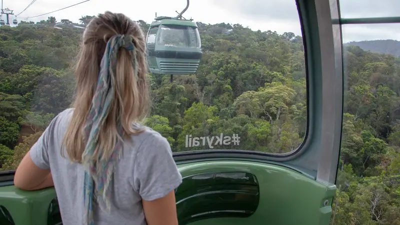 A woman with a ponytail rides the colourful Skyrail cable car high above lush rainforest, looking out at another carriage in the distance.