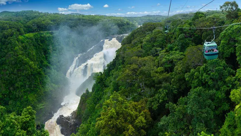 Experience a Skyrail cable car soaring above vibrant rainforest and a stunning waterfall beneath clear blue skies on the way to Rainforestation.
