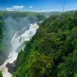 Skyrail cable car travels above vibrant rainforest canopy, overlooking a majestic waterfall under a bright blue sky with fluffy clouds.