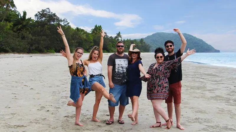 Six smiling holidaymakers on Cape Tribulation's sandy beach, lush Daintree rainforest and mountains behind—Daintree Wilderness Tour scene.