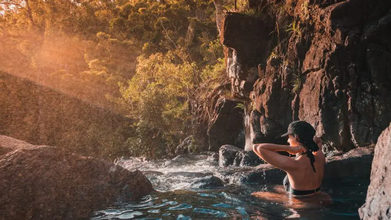 Swimmer unwinds in a crystal-clear rocky pool beneath Falls To Paradise Waterfall, with sunlight streaming through lush forest trees.