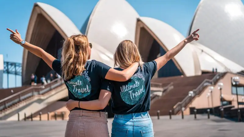 Two joyful women in Ultimate Travel shirts raise their arms, celebrating the guided 7-week East Coast adventure from Melbourne to Cairns.