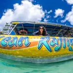 Happy woman leans over vibrant Ocean Rafting Northern Exposure Tour boat in crystal-clear water beneath a bright, blue sky with clouds.