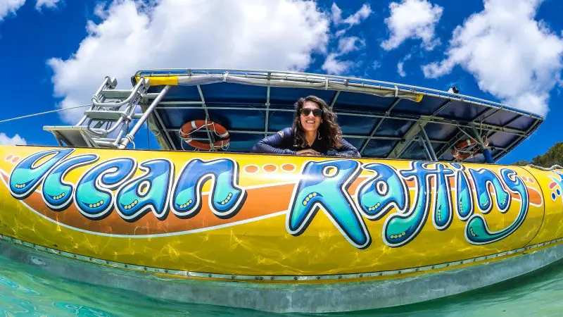 Happy woman leans over vibrant Ocean Rafting Northern Exposure Tour boat in crystal-clear water beneath a bright, blue sky with clouds.
