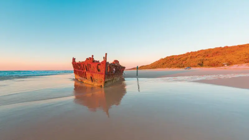 Discover the iconic rusty shipwreck on Fraser Island’s sandy beach at sunset, with gentle waves, lush greenery—Fraser Experience Tour.