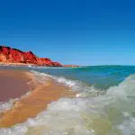 Powerful waves crash onto a pristine sandy beach bordered by striking red rocky cliffs on the 10 Day Broome to Perth Adventure.