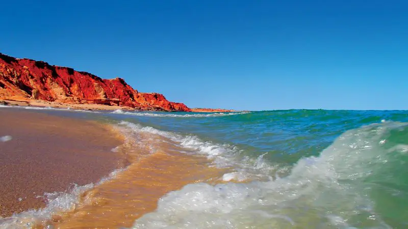 Powerful waves crash onto a pristine sandy beach bordered by striking red rocky cliffs on the 10 Day Broome to Perth Adventure.