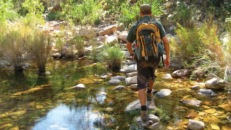 Hiker with rucksack crosses rocky stream on 10 Day Broome to Perth West Coast Adventure, showcasing scenic wilderness and outdoor exploration.