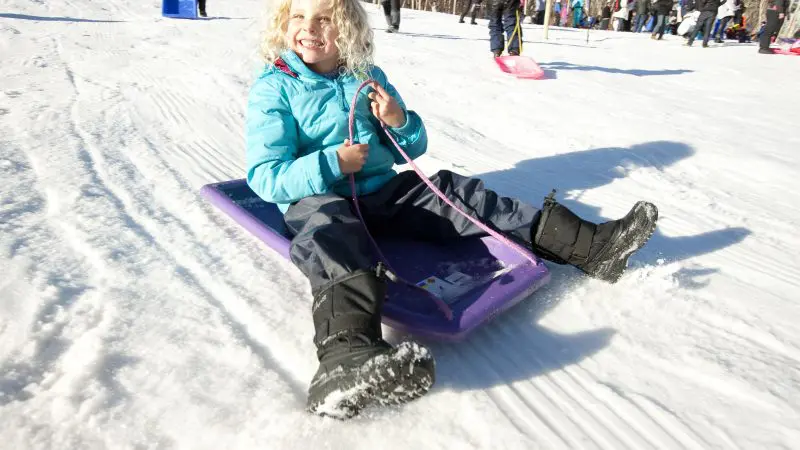 Happy child in a vibrant blue coat rides a purple sledge on snowy Lake Mountain during a 1 Day Snow Tour from Melbourne.