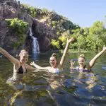 Three joyful women raise their arms and smile whilst swimming under a scenic waterfall on a 1 Day Litchfield National Park tour.
