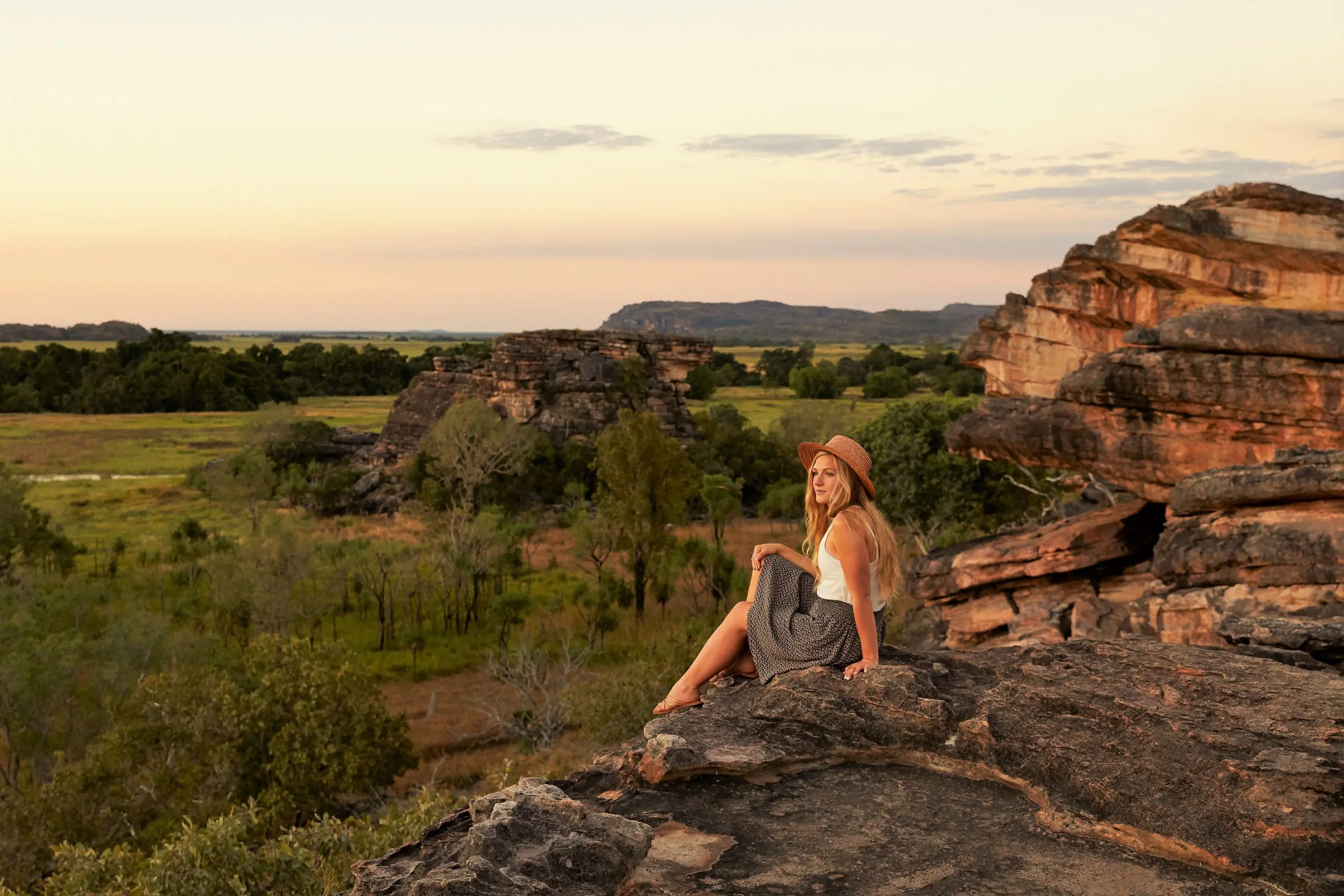 Woman wearing a sunhat admires vibrant Kakadu sunset from a scenic rocky ledge on her 1 Day Wilderness Escape with Crocodile Cruise.