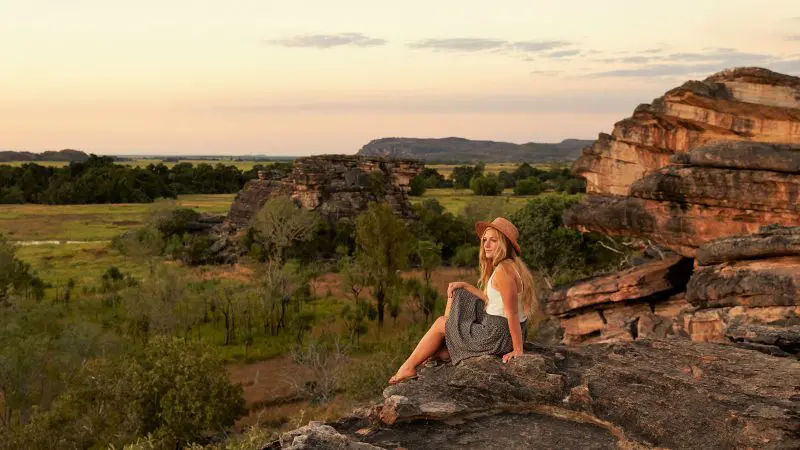 Woman wearing a sunhat admires vibrant Kakadu sunset from a scenic rocky ledge on her 1 Day Wilderness Escape with Crocodile Cruise.