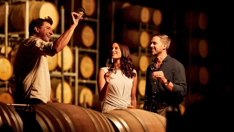 Three people savour wine by oak barrels during a 1 Day Hunter Valley Scenic Wine Tour inside an atmospheric, softly lit winery.