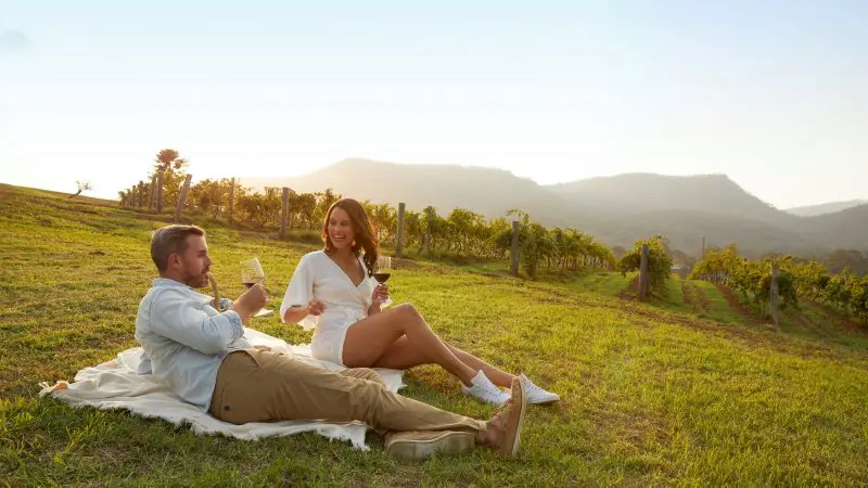 Couple enjoying premium wine on a scenic Hunter Valley tour, relaxing on a blanket with sweeping mountain views at sunset.