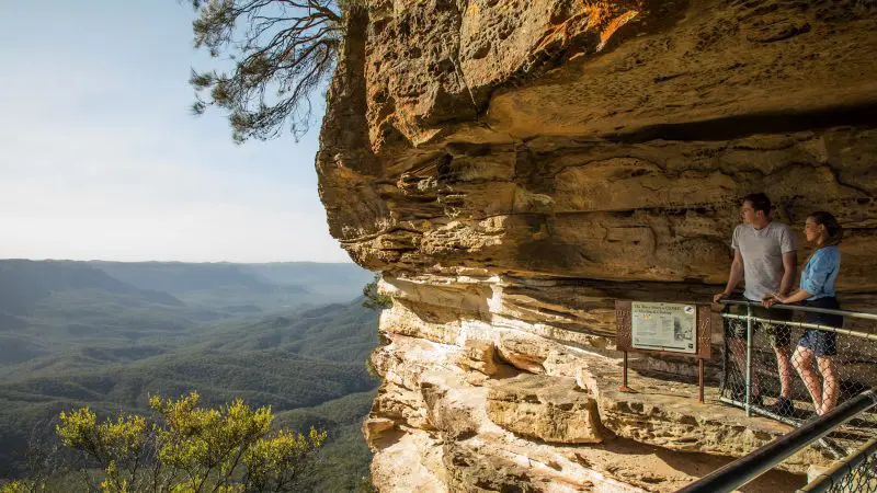Two tourists admire a breathtaking sunset over Blue Mountains wilderness, overlooking lush forest valleys and dramatic mountain scenery.