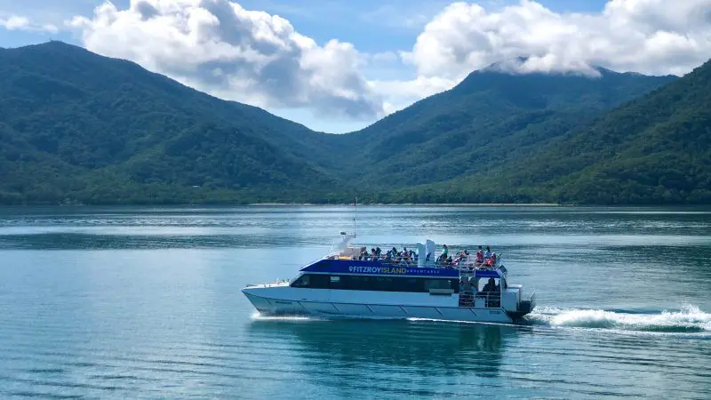 A scenic ferry carrying passengers glides across a tranquil lake, surrounded by majestic mountains and drifting clouds, like a Fitzroy Island Half Day Tour.