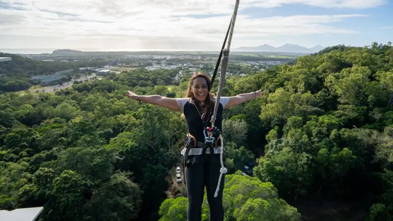 Smiling woman in a safety harness stands with arms outstretched on Walk The Plank Skypark, high above a vibrant green landscape.