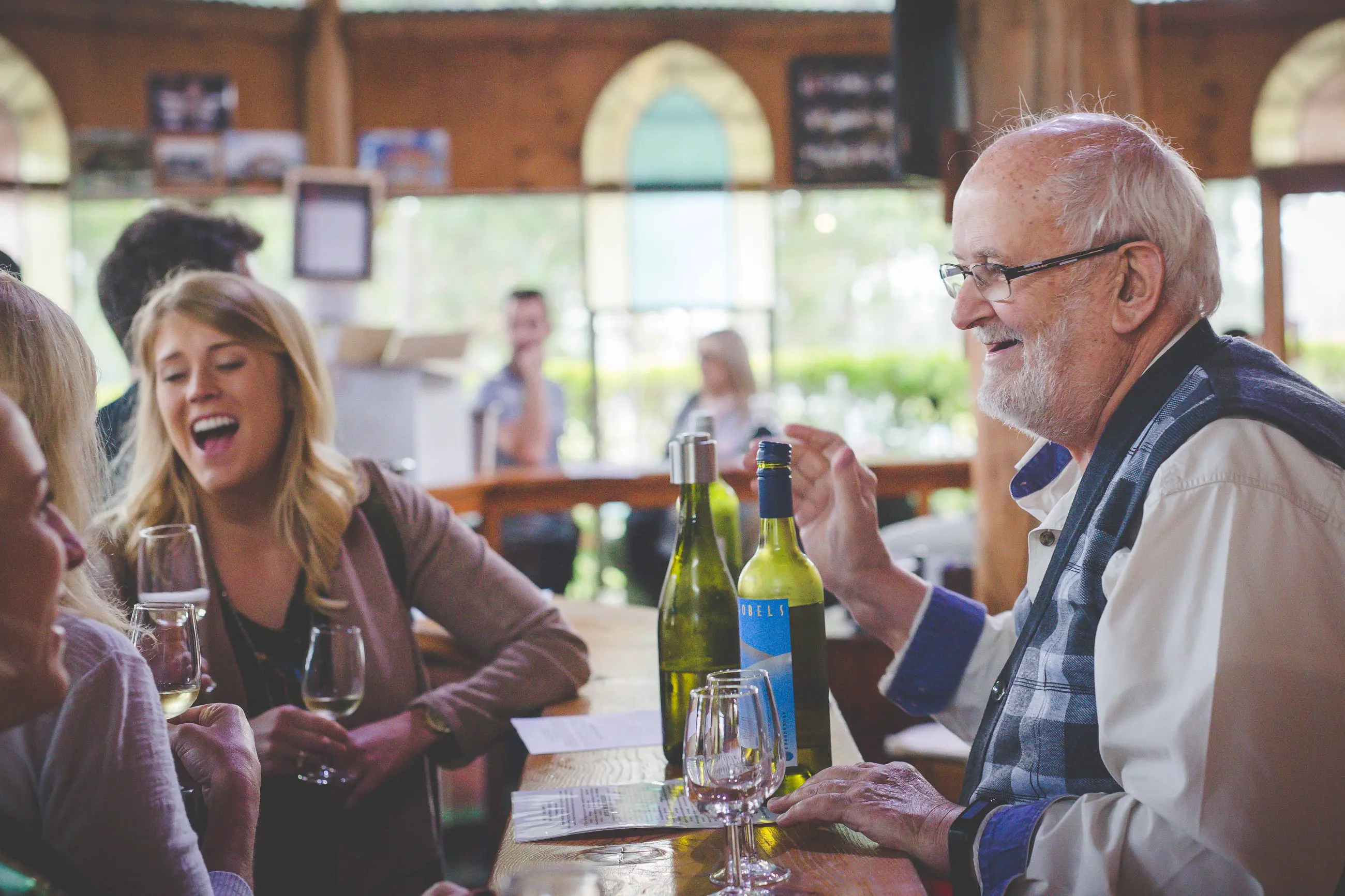 An older man and young woman share laughter during a Hunter Valley Wine Tour in a cosy, sunlit restaurant, enjoying their experience.