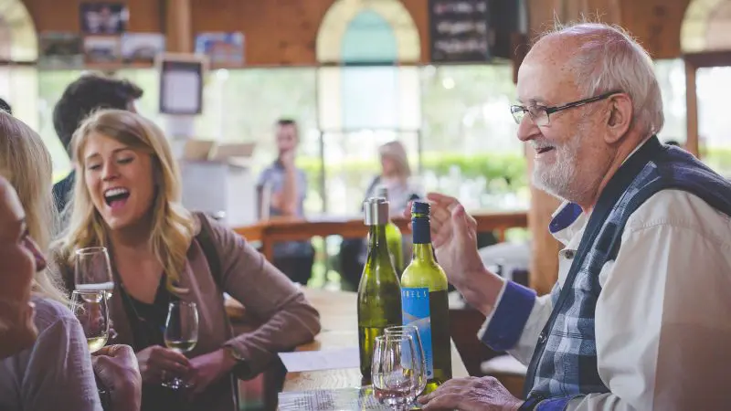 An older man and young woman share laughter during a Hunter Valley Wine Tour in a cosy, sunlit restaurant, enjoying their experience.
