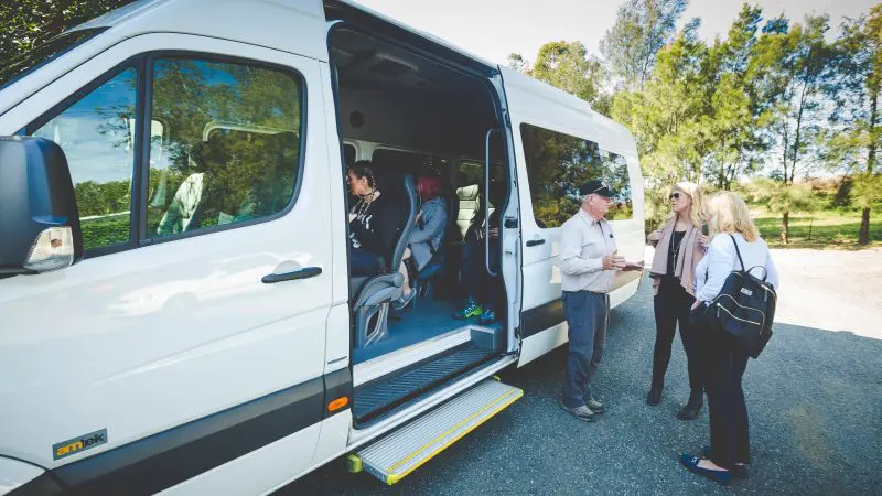 Friends enjoy lively conversation beside a white van with its door open on a sunny Hunter Valley wine tour, perfect for day trips.
