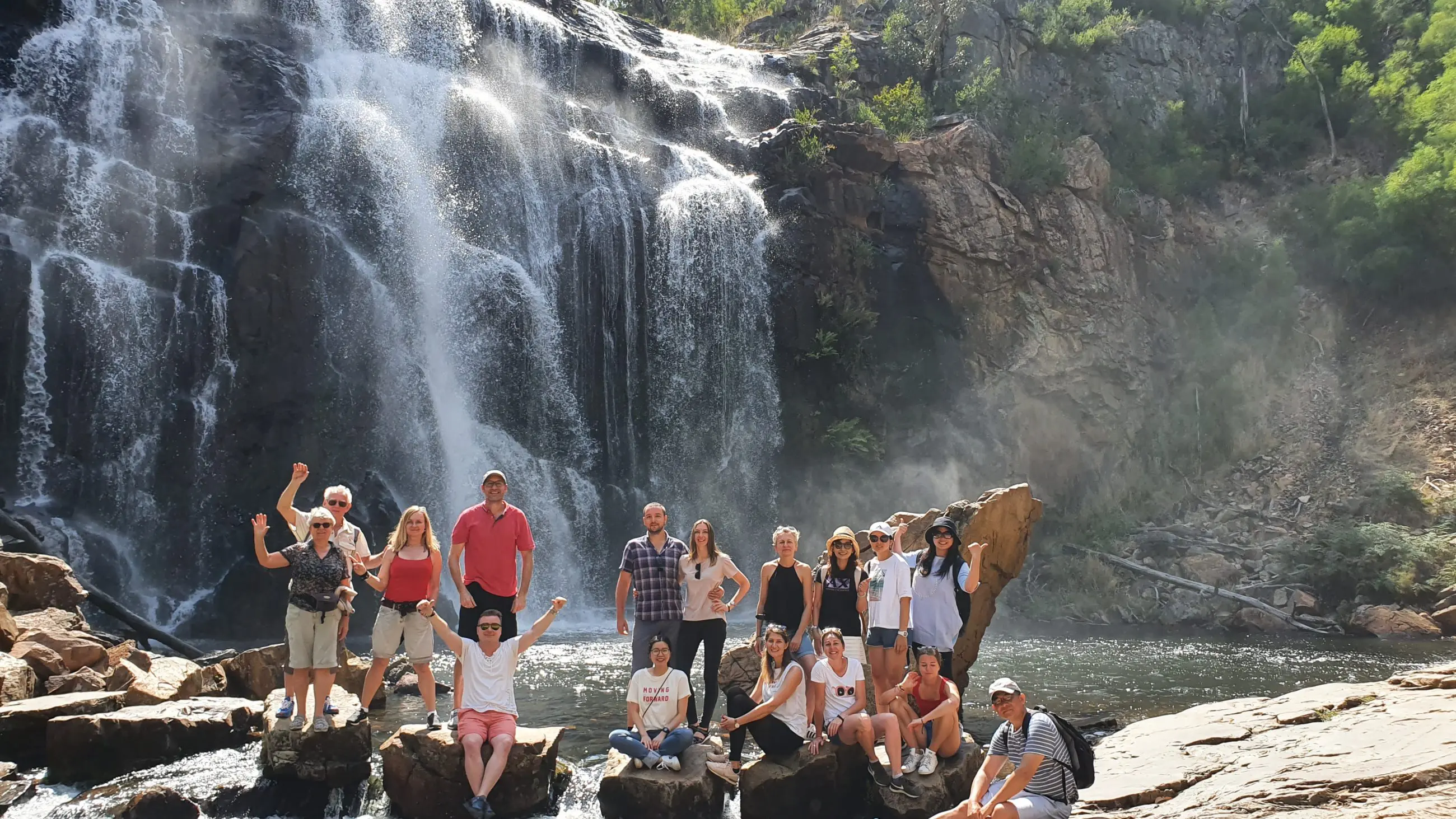 A group enjoys scenic rocks by a majestic Grampians waterfall, framed by lush trees and rugged cliffs on their day tour adventure.
