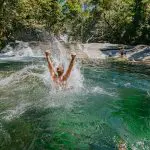 Adventurer joyfully splashes into crystal-clear green river beneath lush Daintree Rainforest canopy, with scenic waterfall behind.