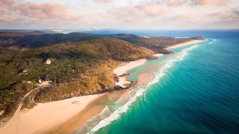 Stunning aerial shot of Fraser Island’s pristine sandy beach, crystal-clear turquoise waters, and lush green hills—ideal for a 2 Day Tag Along Adventure.