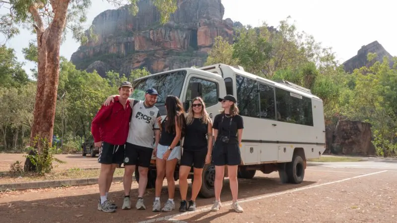 Five happy travellers pose by an off-road coach on a 3 Day Kakadu Adventure, with scenic rocky cliffs and lush trees in the background.