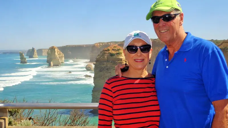 Happy couple wearing sunglasses stands by the sea with scenic rocky cliffs in view on a 1 Day Great Ocean Road Rainforest Tour.