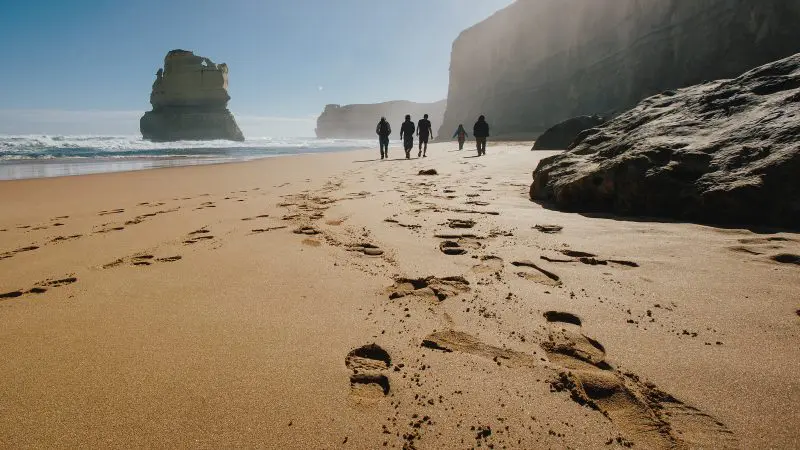 Silhouetted tourists walk along a scenic sandy beach beside rugged cliffs on a 1 Day Great Ocean Road Reverse Eco Tour in Australia.