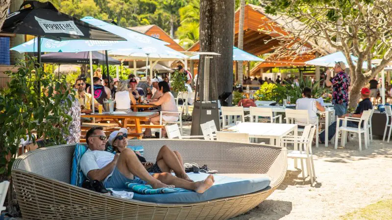 Couple lounging by the vibrant Falls To Paradise Waterfall and Oceanfront Beach Bar, surrounded by lush palm trees and tropical ambience.