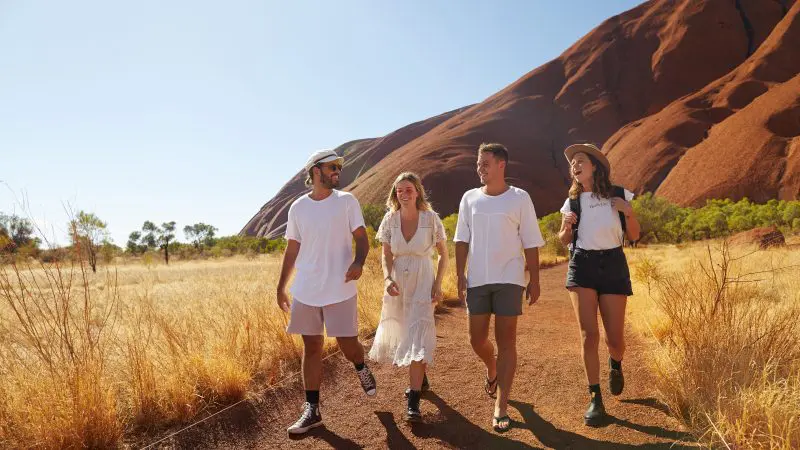 Four travellers hike a scenic dirt track on a 2 Day Uluru Kata Tjuta Rock Tour from Ayers Rock Yulara, iconic red rock formations visible.