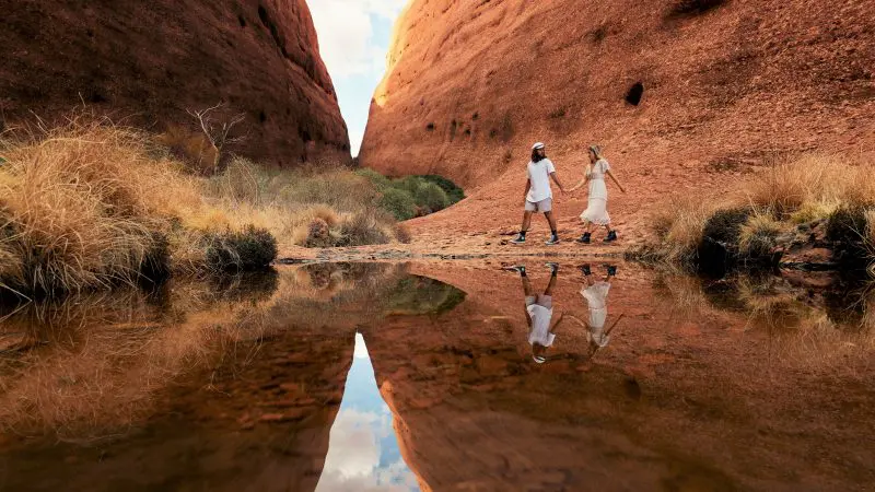 Walkers trek between striking red rocks mirrored in tranquil water, showcasing the unforgettable 3 Day Red Centre Uluru Kings Canyon Safari.