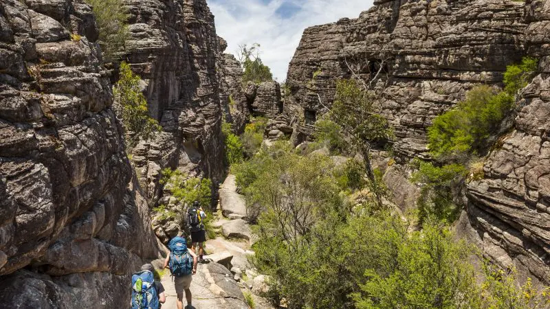 Adventurous hikers with rucksacks trek a scenic, rocky path on a 1 Day Grampians Eco Wilderness Escape amid cliffs and lush greenery.