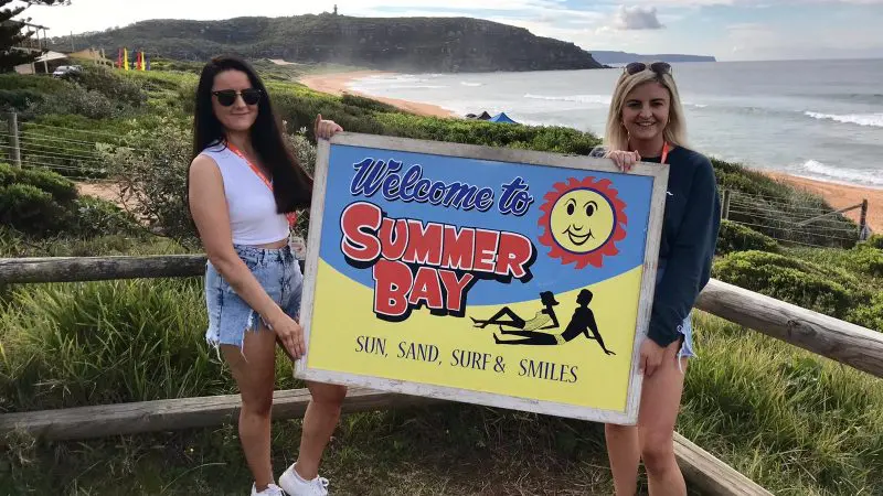 Two smiling women in summer attire hold a Welcome to Summer Bay sign, ideal for your 1 Day Home Away Location Tour experience.