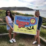 Two women on a sunlit Home Away Tour beach hold a “Welcome to Summer Bay” sign, with lush green hills and blue sea in view.