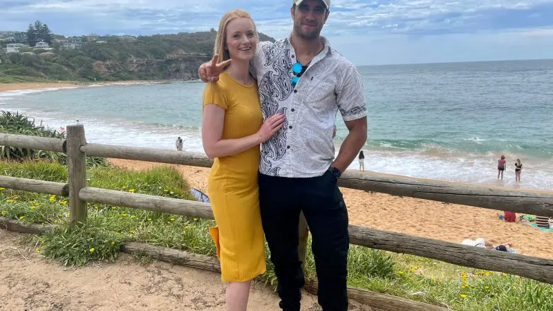Smiling couple on sandy beach path by the sea, enjoying 1 Day Home Away Location Tour under a cloudy sky, perfect holiday getaway.