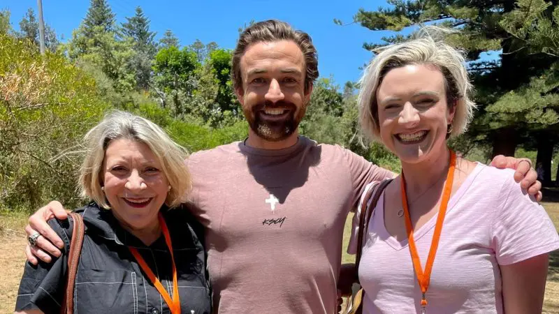 Three adults smiling outdoors wearing Celebtime Tours orange lanyards, enjoying a 1 Day Home Away Tour beneath a clear blue sky.